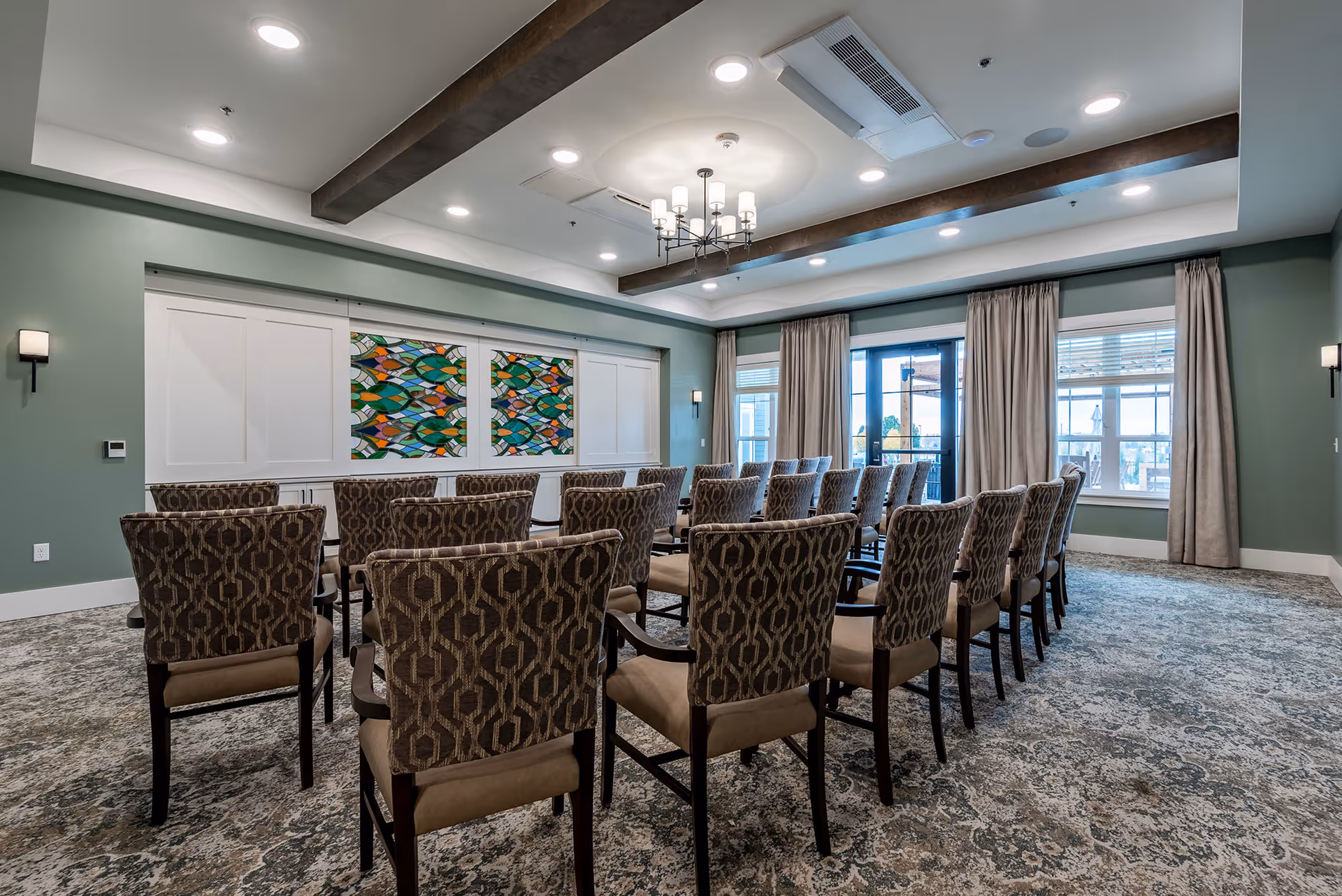 A bright community meeting room with rows of patterned upholstered chairs facing a front wall with colorful artwork, large windows, drapes, and ceiling light fixtures.