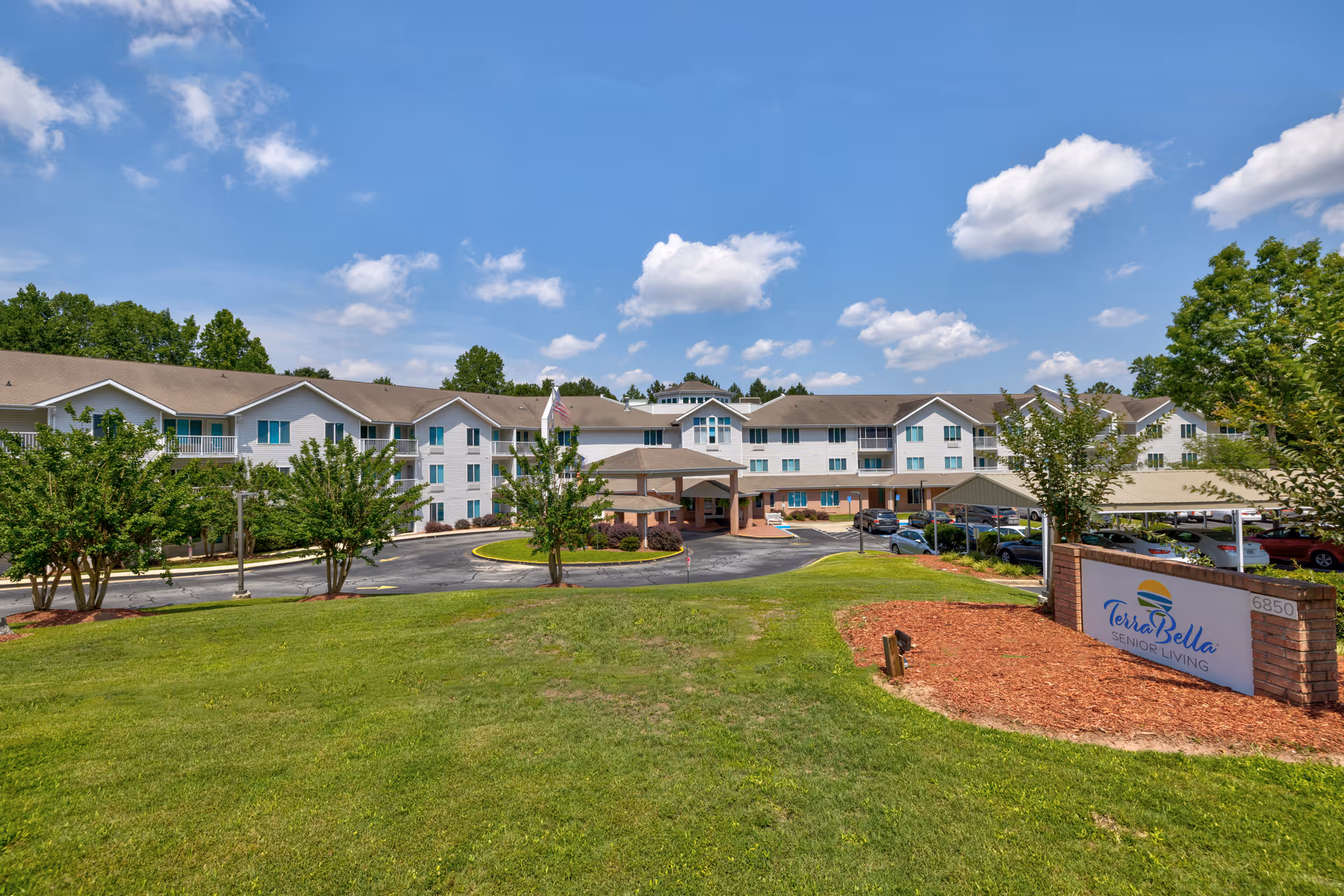 Exterior view of TerraBella Riverplace senior living facility showing a large three-story building with multiple windows and a covered entrance. The building is surrounded by green grass, trees, and a parking area with cars. A sign with the facility name is visible in the foreground on the right side.