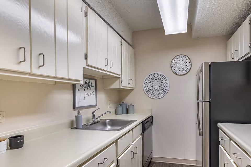 A clean, modern kitchen with white cabinets, a stainless steel sink, dishwasher, and refrigerator. The countertop is light-colored and there are decorative wall hangings and a framed picture with the word 'grace' above the sink.