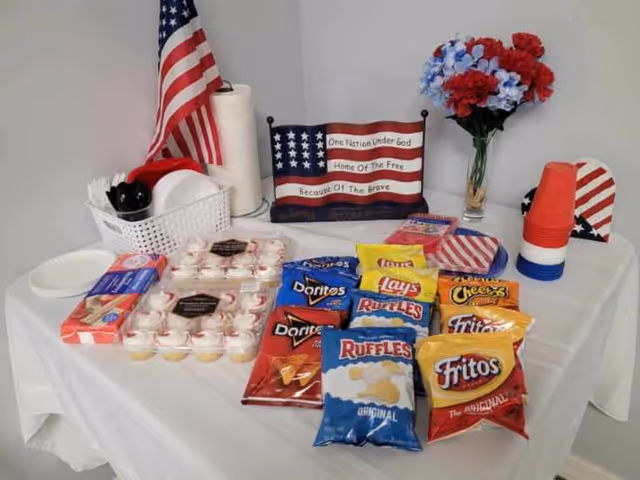 A table covered with a white tablecloth displaying an assortment of snack bags including Doritos, Lay's, Ruffles, Cheetos, and Fritos. There are also cupcakes, red, white, and blue plastic cups stacked, an American flag, a paper towel roll, a basket with plates and utensils, a patriotic sign with the text 'One Nation Under God Home Of The Free Because Of The Brave,' and a vase with red, white, and blue flowers.