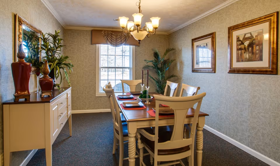 A well-lit dining room with a wooden table set for six, featuring placemats and candles. The room has a window with blinds and a valance, two framed paintings on the wall, a chandelier overhead, and a sideboard with decorative vases and plants.