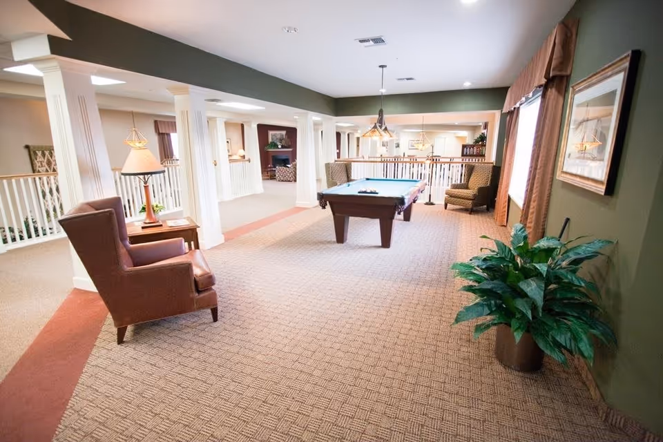 Interior view of a senior living facility lounge area featuring a pool table in the center, surrounded by comfortable armchairs, a side table with a lamp, and a large potted plant. The room has carpeted floors, green walls, and large windows with curtains allowing natural light to enter.