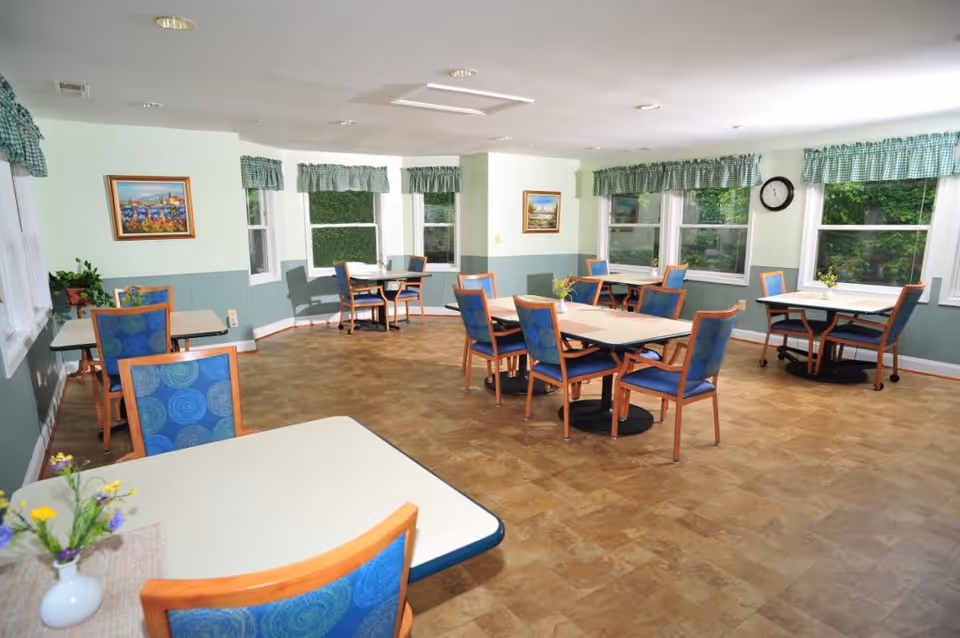 A bright dining room with multiple tables and blue cushioned chairs arranged neatly. The room has large windows with green checkered valances, light green walls with a darker green wainscoting, and framed paintings on the walls. Small flower arrangements are placed on the tables, and a clock is mounted on one wall.