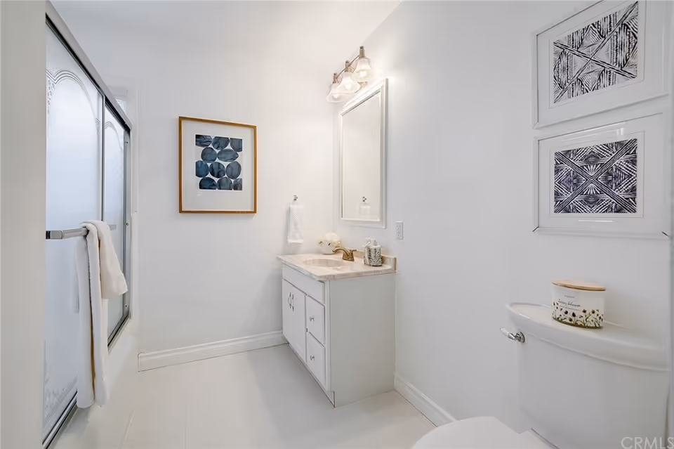 A clean, white bathroom featuring a shower with frosted glass sliding doors and a white towel hanging on the handle. There is a white vanity with a marble countertop, a sink, a mirror above it, and a light fixture with three bulbs. On the right side, there is a toilet with a decorative container on top and two framed abstract artworks hanging on the wall above it.