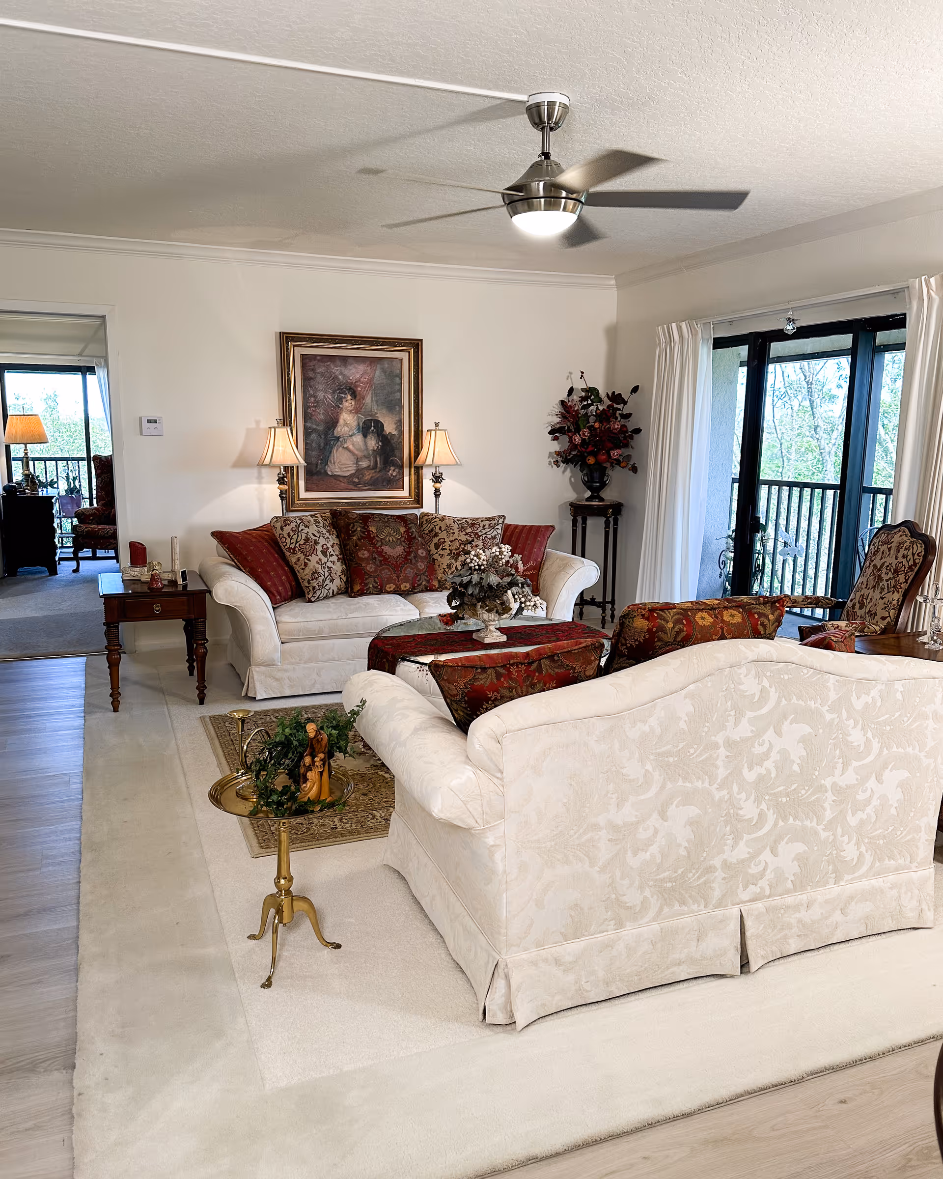 A cozy living room with two cream-colored sofas adorned with patterned cushions, a small brass side table with a decorative figurine, a coffee table with a floral centerpiece, and a framed painting on the wall. The room has a ceiling fan with light, a side table with two lamps, and large windows with white curtains leading to a balcony with a view of trees.
