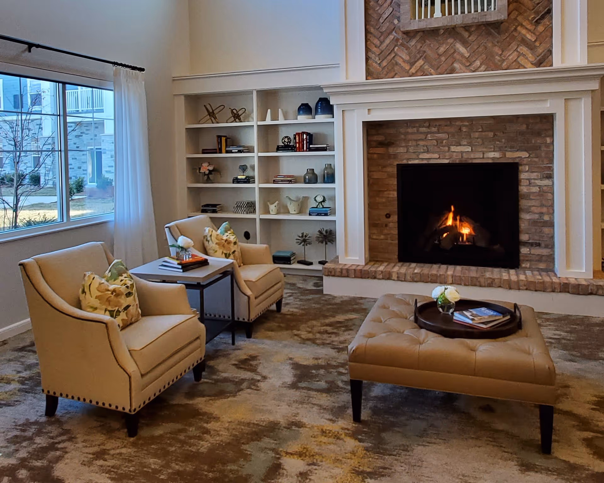 A cozy living room with two beige armchairs featuring floral pillows, a tufted ottoman with a tray holding a small flower vase and books, a brick fireplace with a fire burning, and built-in white shelves decorated with books and ornaments. A large window with white curtains lets in natural light.