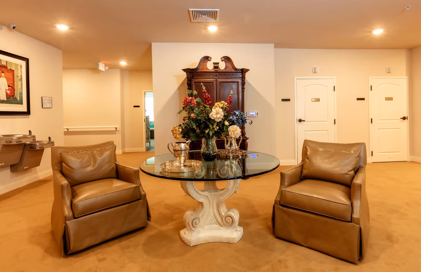 A cozy seating area with two brown leather armchairs facing a round glass table with a decorative white base. On the table is a vase with colorful flowers and silver pitchers on trays. Behind the table is a dark wooden cabinet against a beige wall. The room has beige carpet and walls, with two white doors on the right side and a hallway leading to another room in the background. Two water fountains and a framed picture are visible on the left wall.
