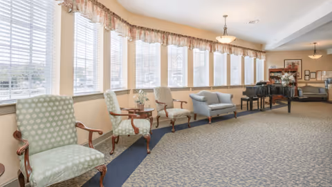 A spacious, well-lit common area with large windows along one wall covered with white blinds and floral valances. The room features several upholstered armchairs and a loveseat arranged along the window side, a black grand piano, a wooden bookshelf with framed pictures and decorative items, and soft beige walls with carpeted flooring.
