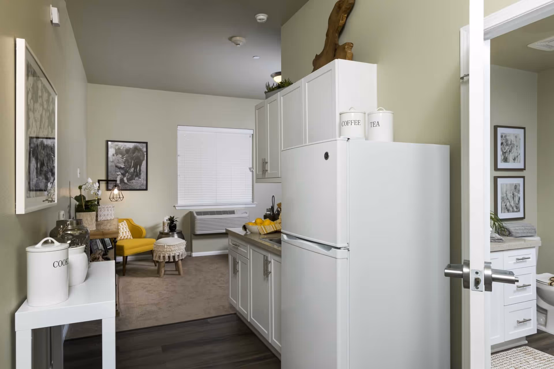 View of a small kitchen area with white cabinets and a white refrigerator. On top of the refrigerator are containers labeled 'COFFEE' and 'TEA'. To the left is a small white table with a container labeled 'COOKIES'. Beyond the kitchen is a living area with a yellow armchair, a small round ottoman, and framed black and white pictures on the walls. To the right, an open door reveals part of a bathroom with white cabinetry and framed pictures on the wall.