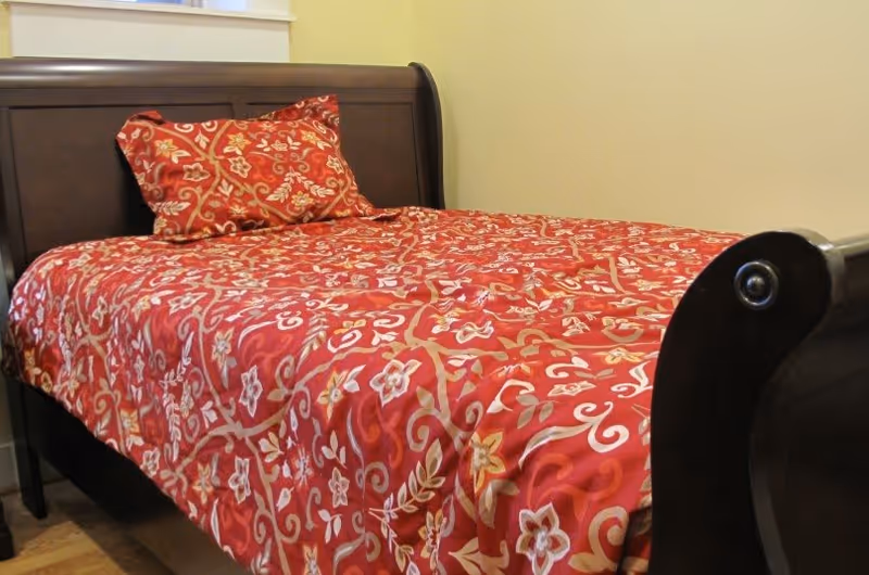 A wooden bed with a dark finish, made up with a red bedspread and matching pillow featuring a floral and vine pattern. The bed is positioned against a light-colored wall with a window above the headboard.