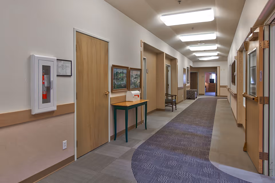 Carpeted interior hallway of a senior living facility with doors, ceiling lights, artwork, and a small table and chair.