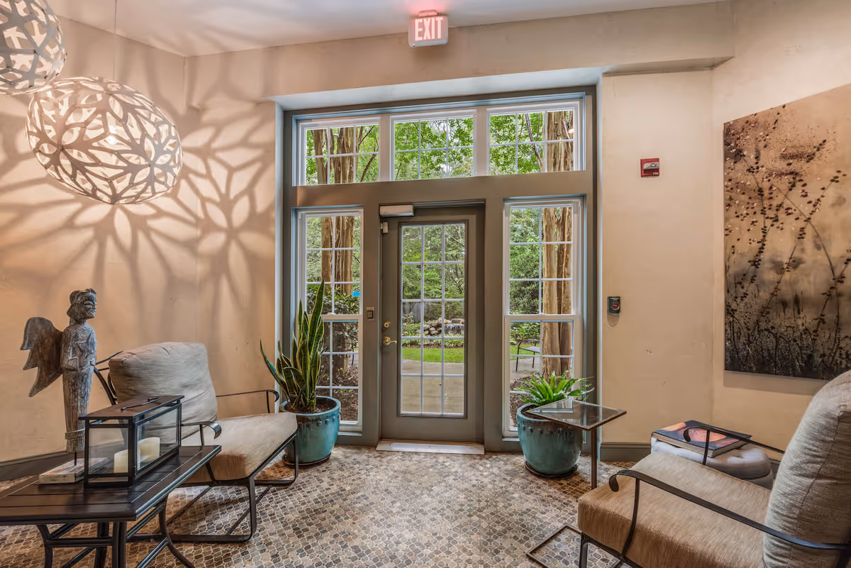 A cozy indoor seating area with two cushioned chairs and a small table between them. The space features a glass door and windows looking out to a green garden. Decorative hanging lights cast intricate shadows on the wall. There are potted plants on either side of the door and a piece of abstract art on the right wall.