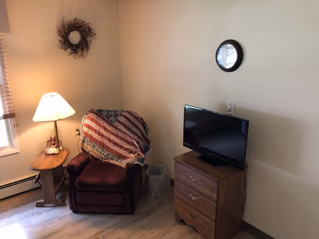 A cozy corner of a room featuring a burgundy recliner chair draped with an American flag-themed blanket, a wooden side table with a lamp and small decorative items, a small wooden dresser with a flat-screen TV on top, a round wall clock, and a decorative wreath hanging on the wall. The room has light-colored walls and wood flooring.