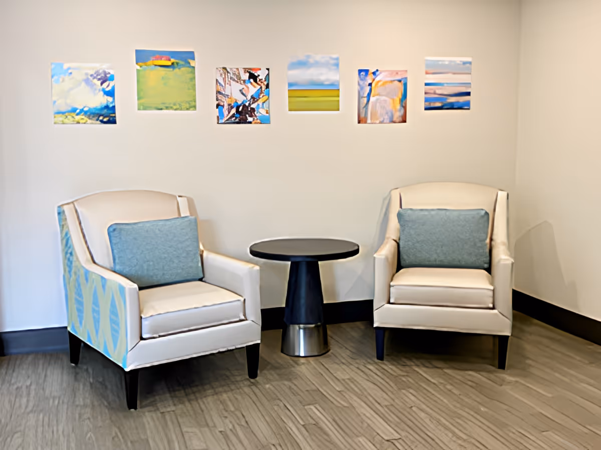 Two upholstered armchairs with blue cushions flank a small round table beneath a row of colorful square paintings on a white wall.