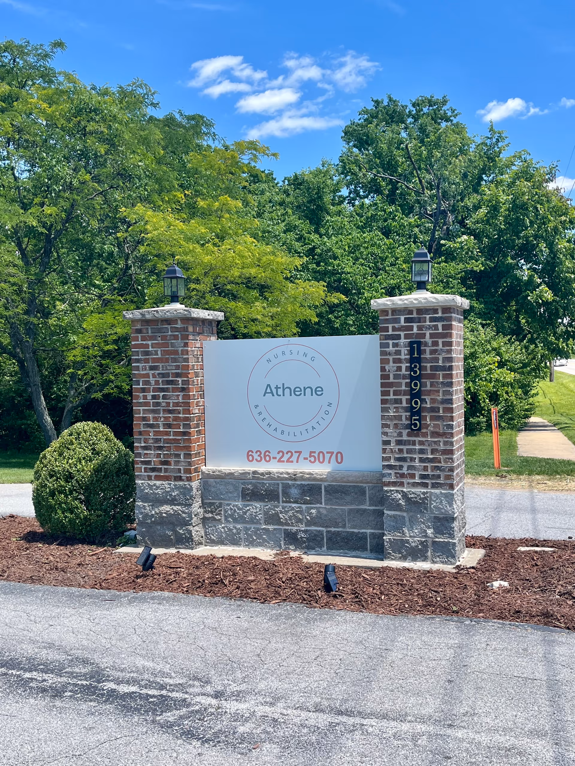 Brick and stone entrance sign for Athene Nursing and Rehabilitation set between two pillars with trees and a blue sky behind it.
