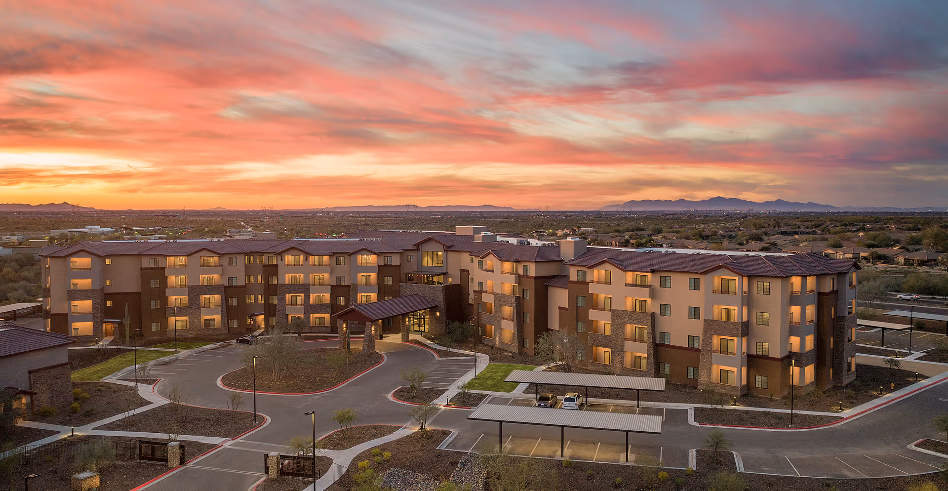 Aerial view of Vista Winds Independent Living facility at sunset, showing a large multi-story building with multiple windows and balconies, surrounded by parking areas and landscaped grounds with pathways and small trees. The sky is vibrant with shades of orange, pink, and purple as the sun sets behind distant mountains.