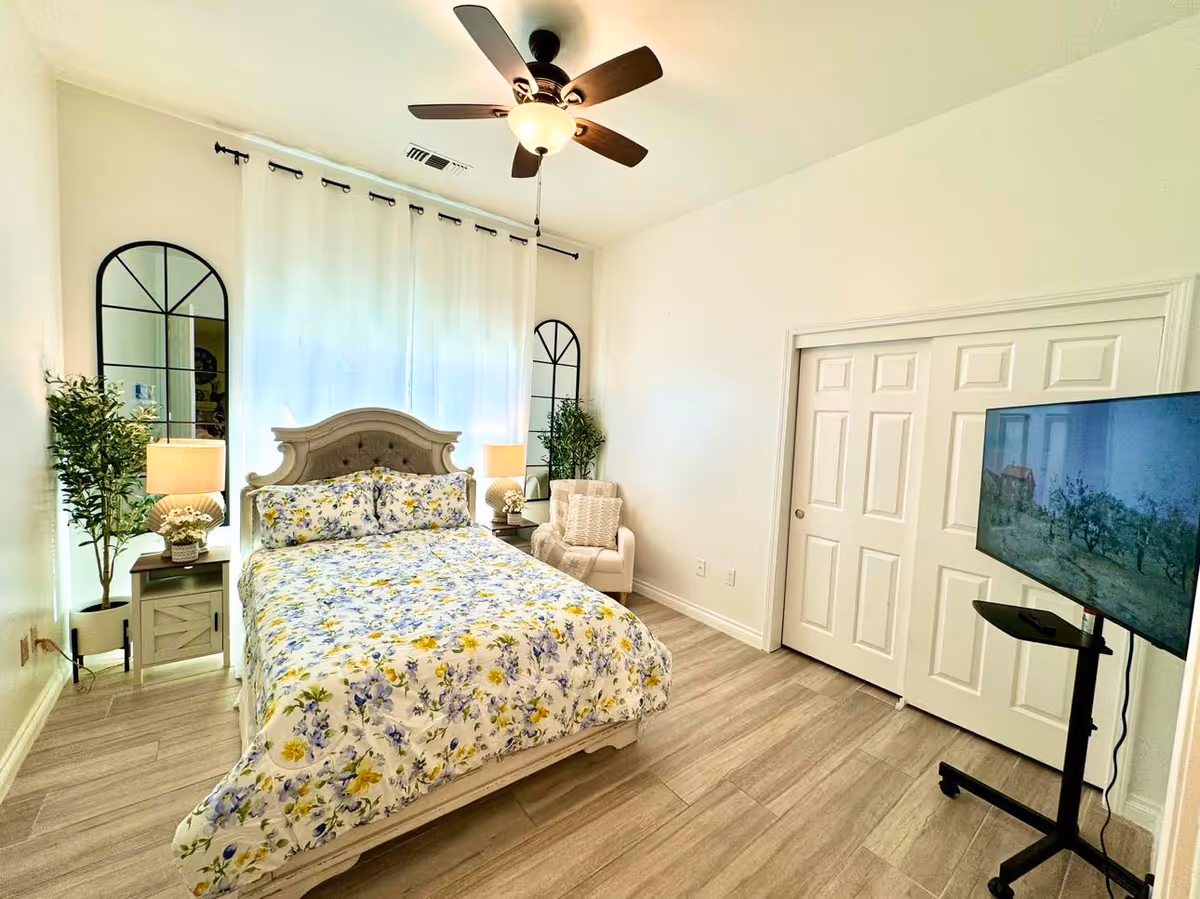 Sunny bedroom featuring a floral-pattern bed with nightstands and lamps, an armchair, large mirrors, a ceiling fan, and a TV on a rolling stand.