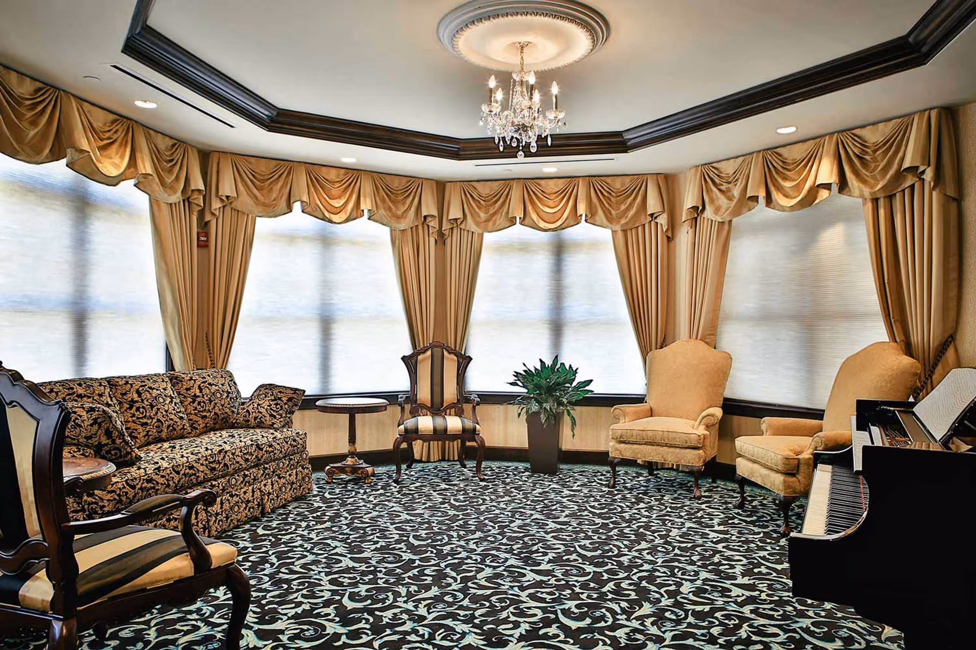 A cozy living room with patterned carpet, a chandelier, large windows with gold drapes, a patterned sofa, several upholstered chairs, a small round table, a potted plant, and a black piano with sheet music.
