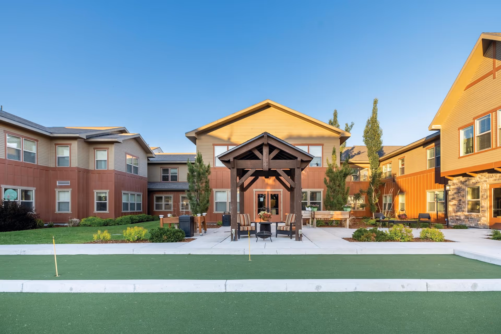 Courtyard with a covered seating pavilion and putting green in front of a multi-story senior living building under a clear blue sky.
