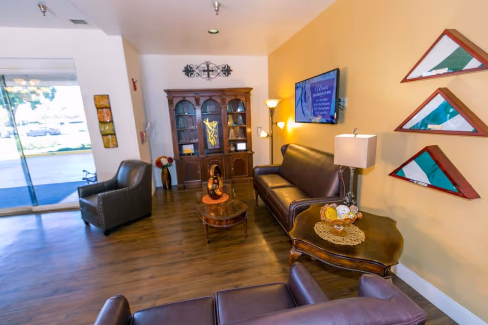A cozy living room area with brown leather sofas and armchairs, a wooden coffee table and side table with decorative items, a wooden cabinet with shelves, a floor lamp, and a wall-mounted TV. The walls are painted yellow and white, with three triangular display cases mounted on one wall. A glass door leads outside, letting in natural light.