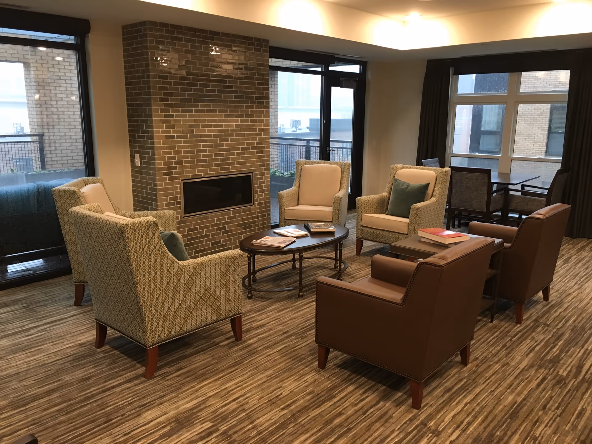 A cozy living room area with six armchairs arranged around a central oval coffee table. The chairs include four fabric-upholstered and two leather-upholstered ones. There is a brick fireplace in the middle of the room, large windows, and a door leading to an outdoor balcony. A dining table with chairs is visible in the background near the windows.