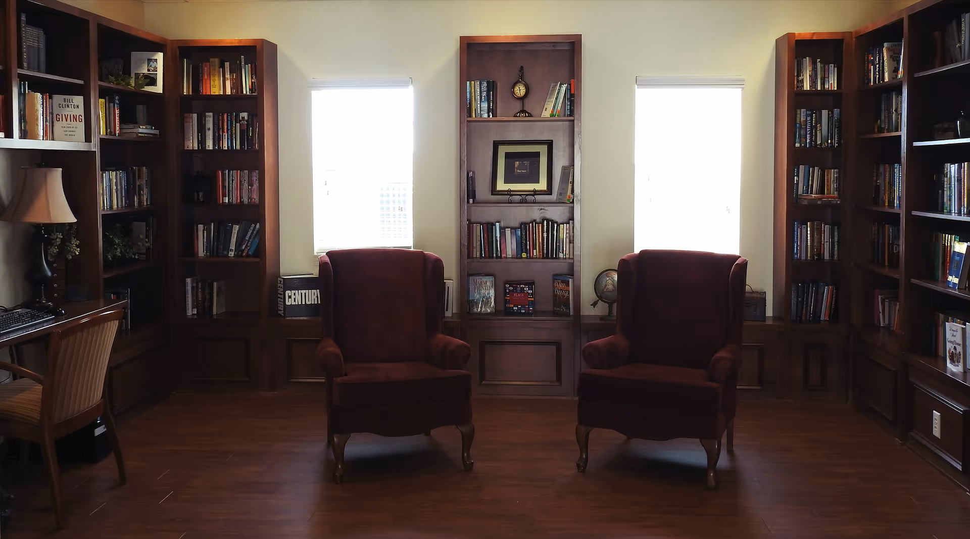 A cozy library or reading room with wooden bookshelves filled with books lining the walls. Two burgundy upholstered armchairs are positioned in front of two windows with blinds, and a wooden desk with a chair and lamp is visible on the left side.