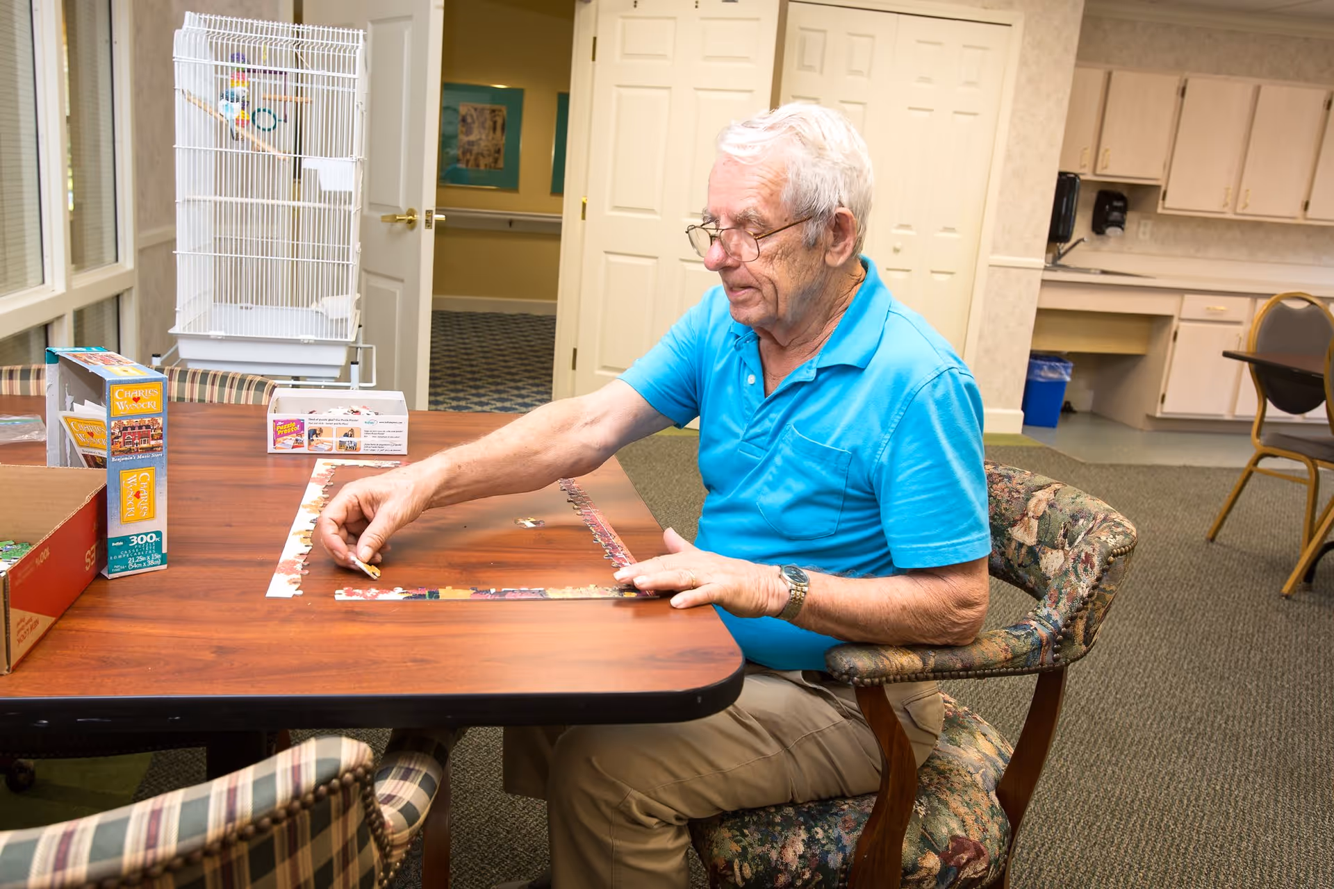 An elderly man wearing glasses and a blue polo shirt is sitting at a wooden table working on a jigsaw puzzle in a room with carpeted floors, floral upholstered chairs, and a white birdcage in the background.