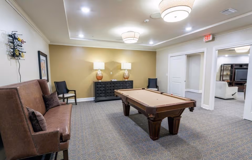 A well-lit recreational room featuring a pool table in the center, a brown leather bench with pillows on the left, two chairs against the far wall, and a black cabinet with two lamps on top. The room has patterned carpet flooring, beige walls, and ceiling lights. An adjacent room with a TV and seating area is visible through an open doorway.