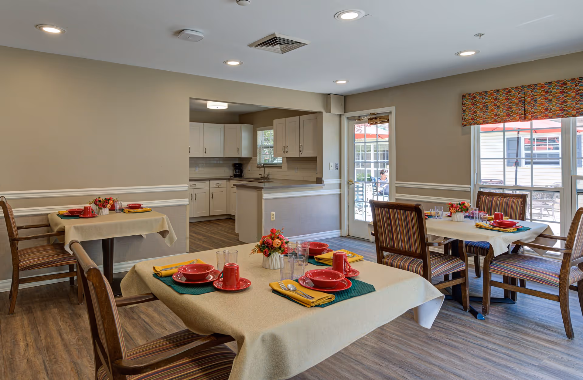 A bright dining room with several tables covered in beige tablecloths, each set with red dishes, cups, yellow napkins, and small flower arrangements. The room has wooden chairs with striped cushions, wood flooring, and beige walls with white trim. In the background, there is a kitchen area with white cabinets and a coffee maker, and a glass door leading to an outdoor patio with tables and red umbrellas.