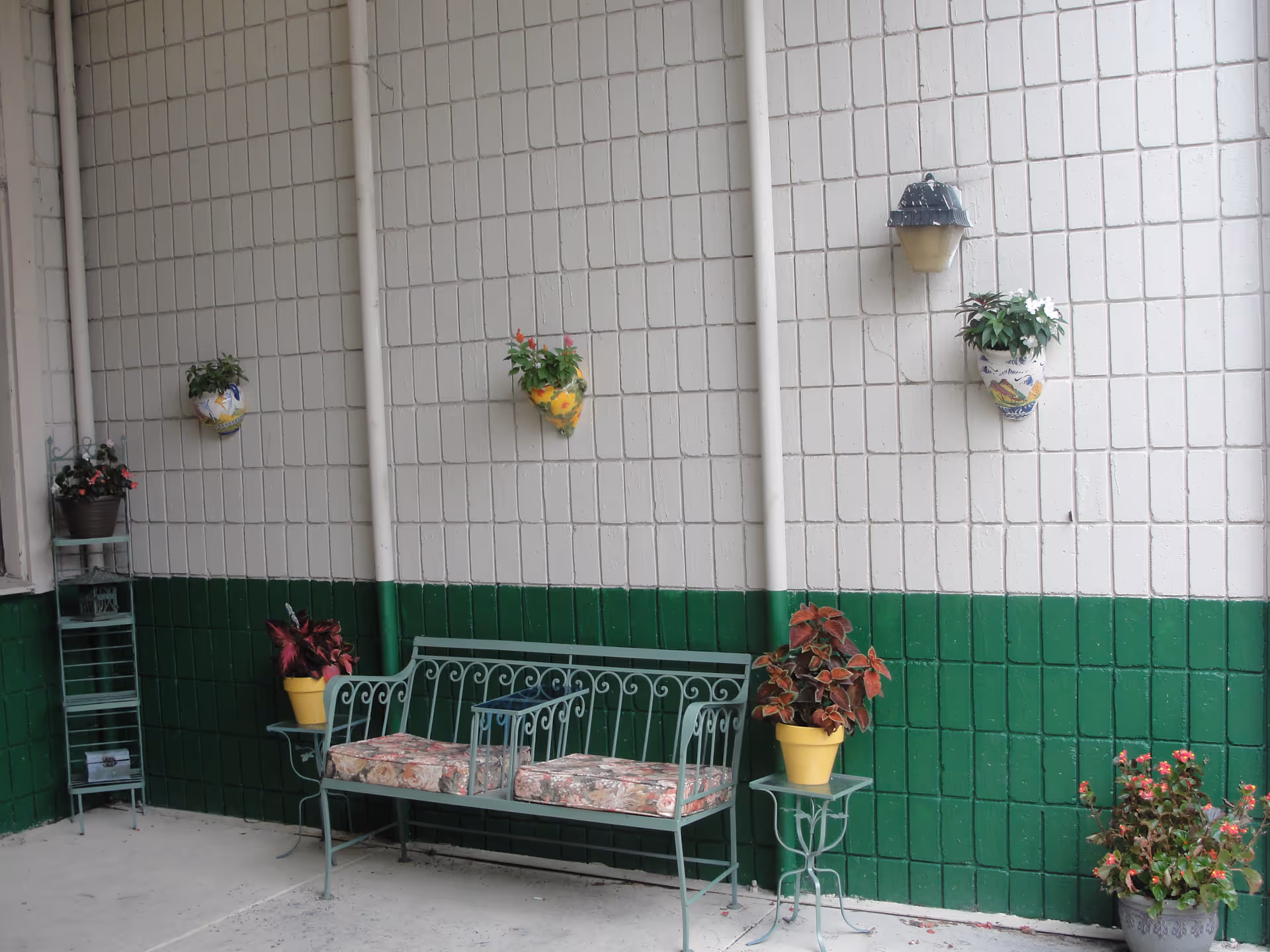 Green metal bench with floral cushions against a white-and-green tiled wall decorated with potted plants and wall planters.