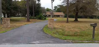 A paved driveway curves toward a single-story building amid trees and grassy lawn.