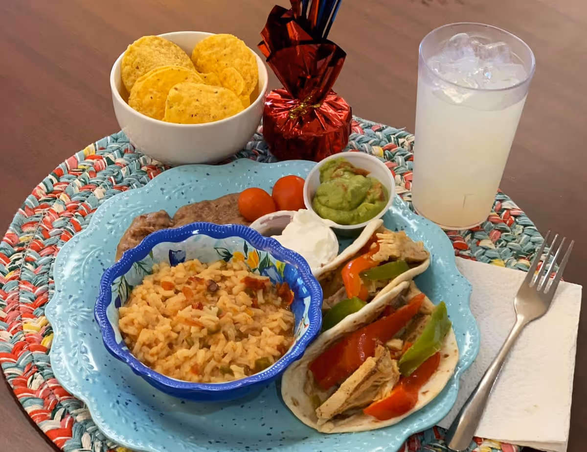 A meal served on a blue plate with two chicken fajitas topped with bell peppers, a small bowl of seasoned rice, refried beans, two cherry tomatoes, a small cup of guacamole, and a small cup of sour cream. There is also a bowl of tortilla chips, a glass of iced beverage, a fork on a napkin, and a colorful woven placemat on a wooden table.