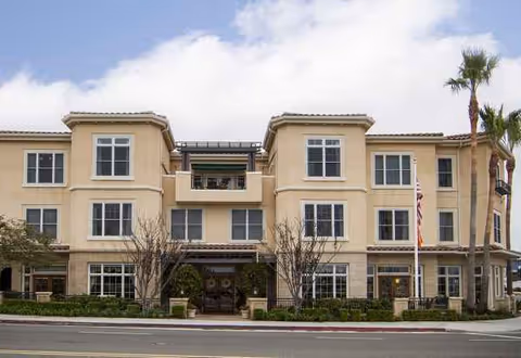 Three-story beige senior living building with entrance, palm trees, and American flag.