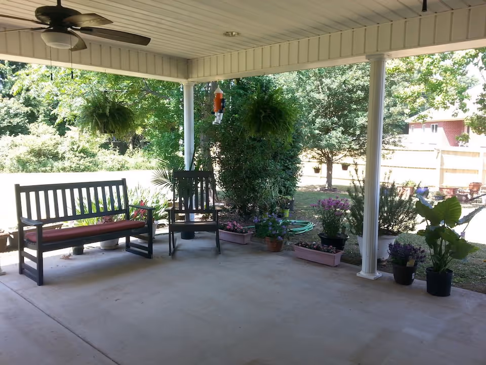 Covered outdoor patio area with a ceiling fan, a bench with a red cushion, a chair, and various potted plants and flowers arranged along the edge. The patio overlooks a green yard with trees and a wooden fence in the background.