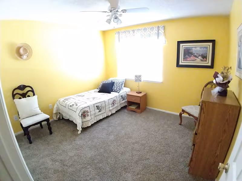 Small bright bedroom with a single bed, nightstand, dresser, chairs, and yellow walls.