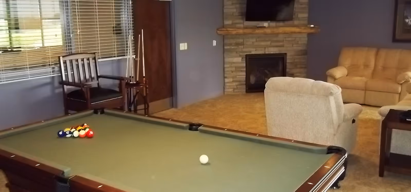 A recreational room featuring a pool table with billiard balls arranged for a game. In the background, there are comfortable beige sofas, a stone fireplace with a mounted TV above it, a wooden chair, and a rack holding pool cues. The room has a cozy and inviting atmosphere with carpeted flooring and windows with blinds.