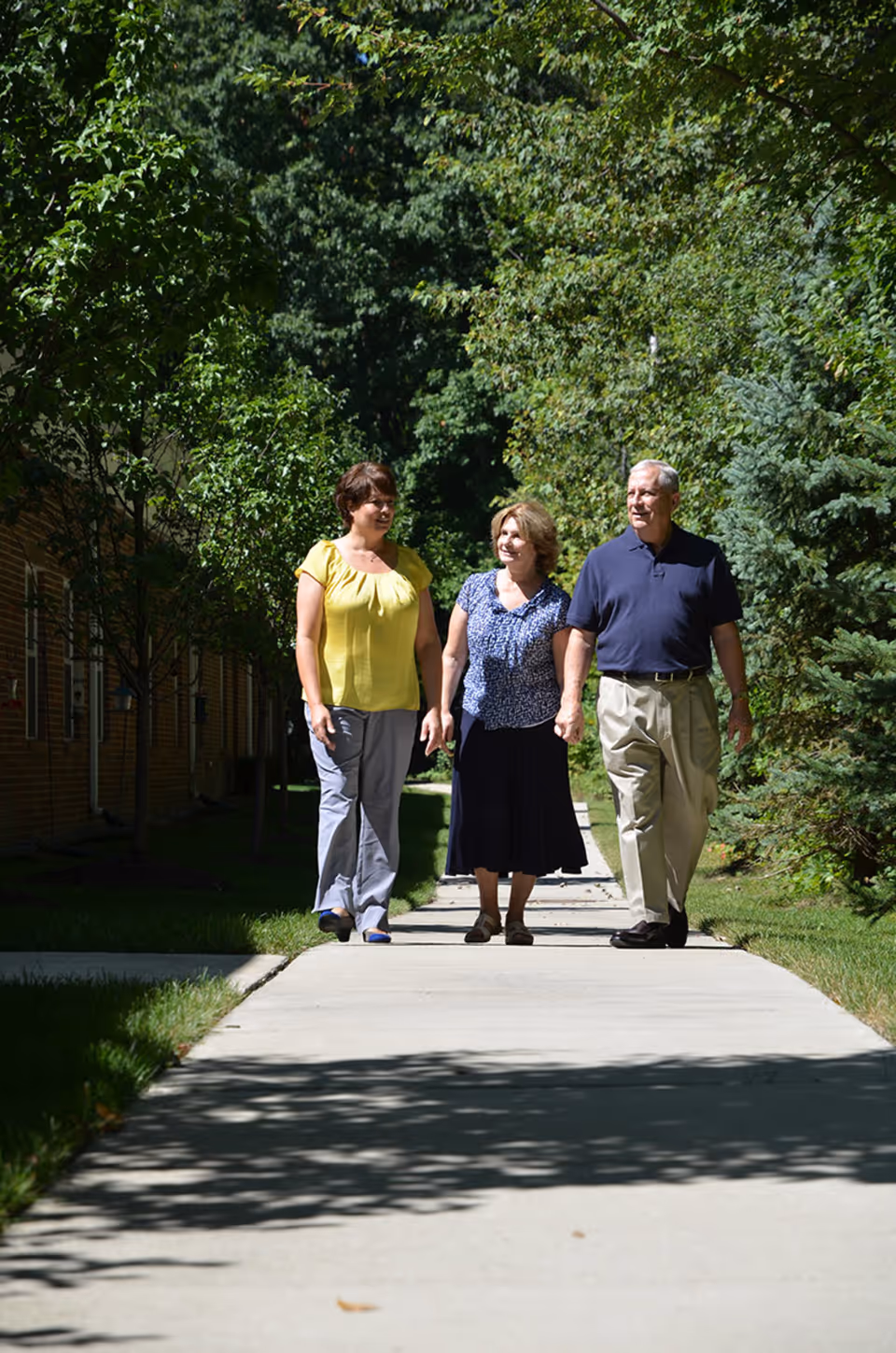 Three adults walking together on a shaded sidewalk surrounded by green trees and grass, with a brick building visible on the left side.