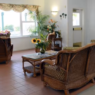 Sunlit communal seating area with wicker chairs around a coffee table, plants, and a window.