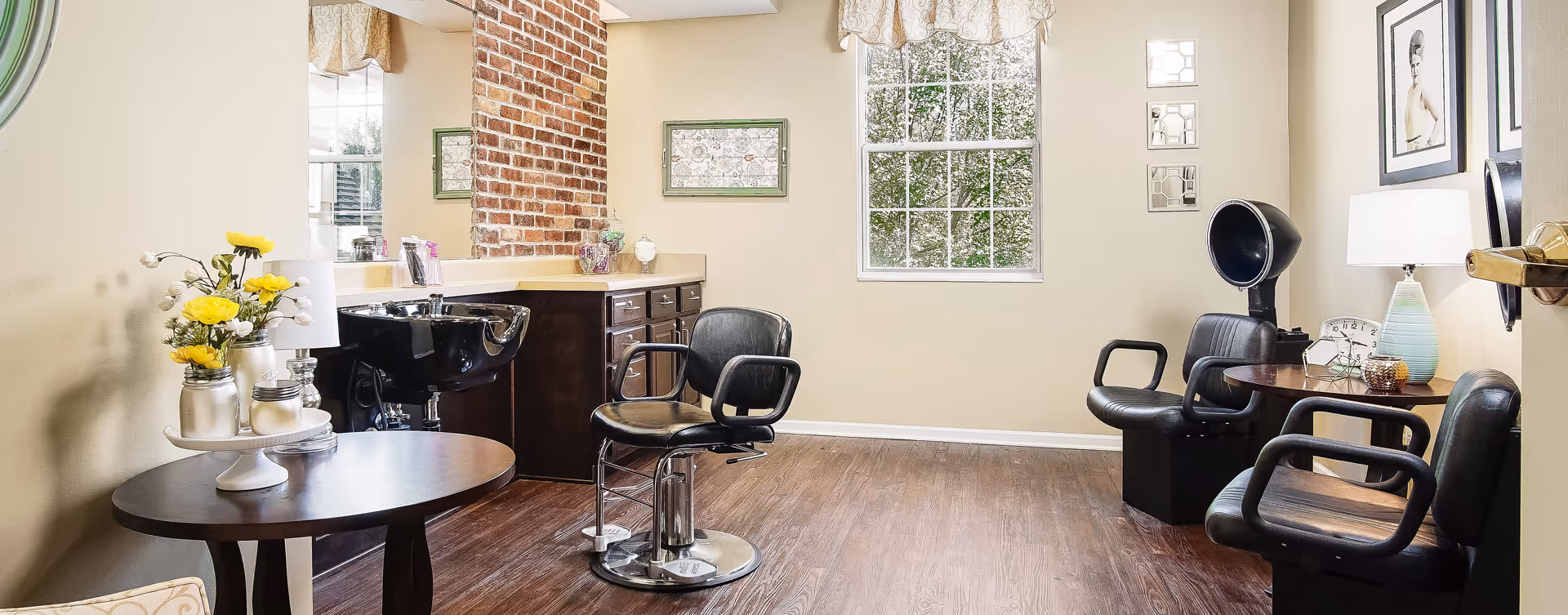 A bright and clean hair salon area with two black salon chairs, a black hair washing sink, wooden cabinets, a large window with a valance, and decorative items including flowers, framed pictures, and a lamp.