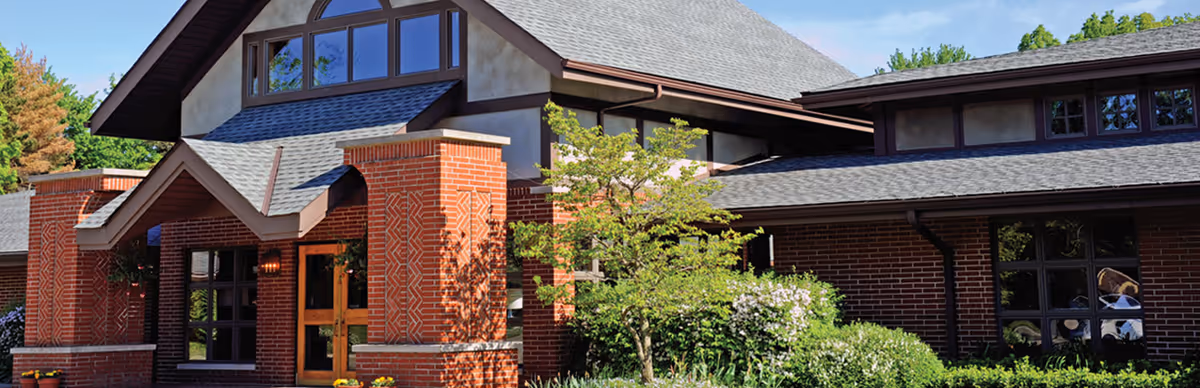 Exterior view of a brick building with large windows, a peaked roof, and a landscaped garden with green bushes and a small tree in front under a blue sky.