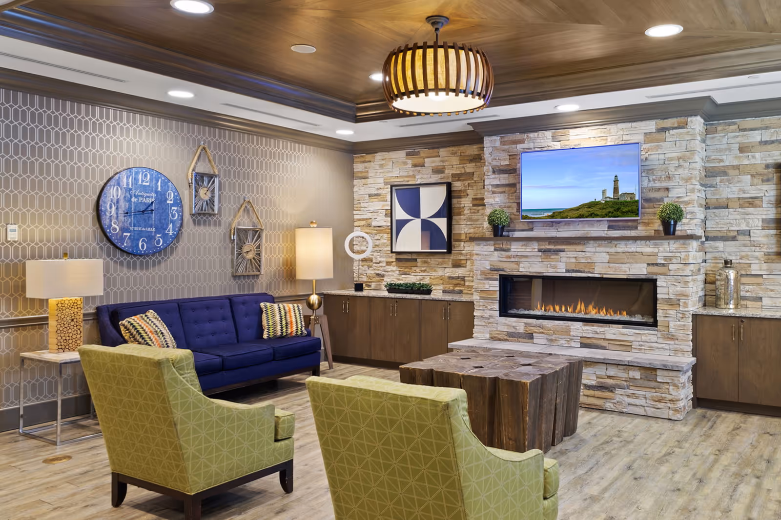 A cozy living room area with a modern stone fireplace and a flat-screen TV mounted above it displaying a lighthouse scene. The room features a blue sofa with patterned pillows, two green armchairs, a wooden coffee table, two floor lamps, and decorative wall art including a large blue clock and geometric designs. The ceiling has a wooden finish with a circular pendant light.