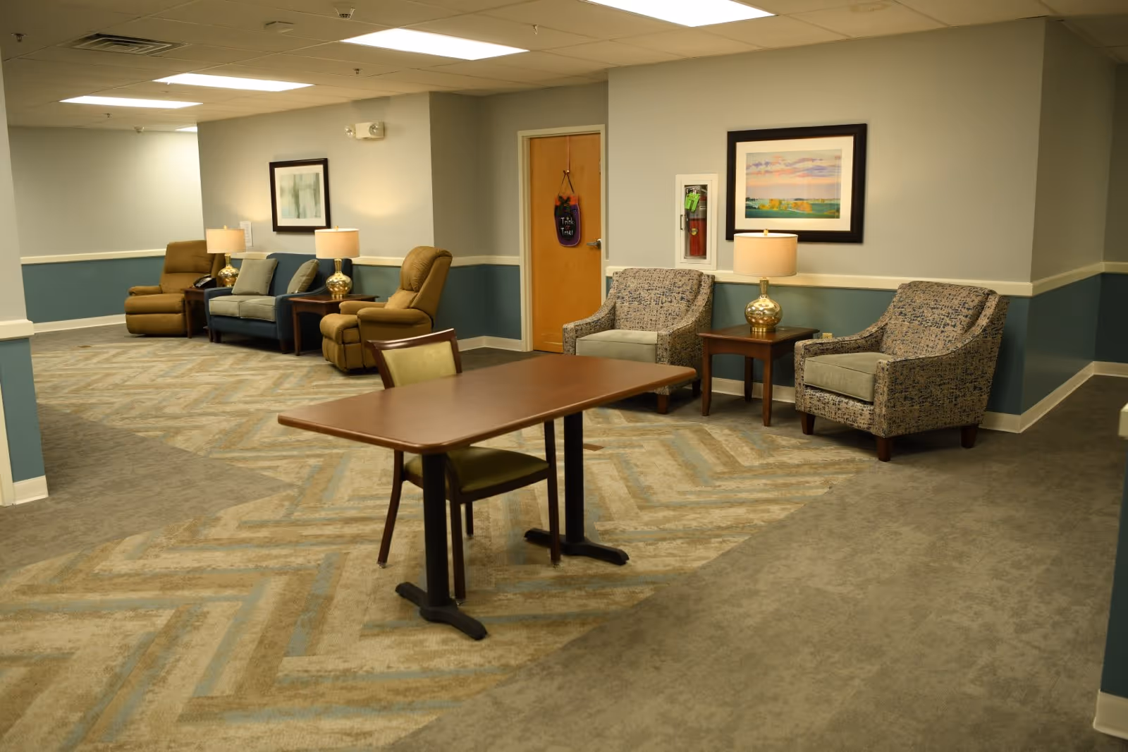 A cozy seating area in a senior living facility hallway with two patterned armchairs and a side table with a lamp, a wooden table with a chair in the foreground, and a sofa with two recliners and lamps along the wall. The walls are painted light gray with a teal wainscoting, and framed artwork hangs above the furniture.