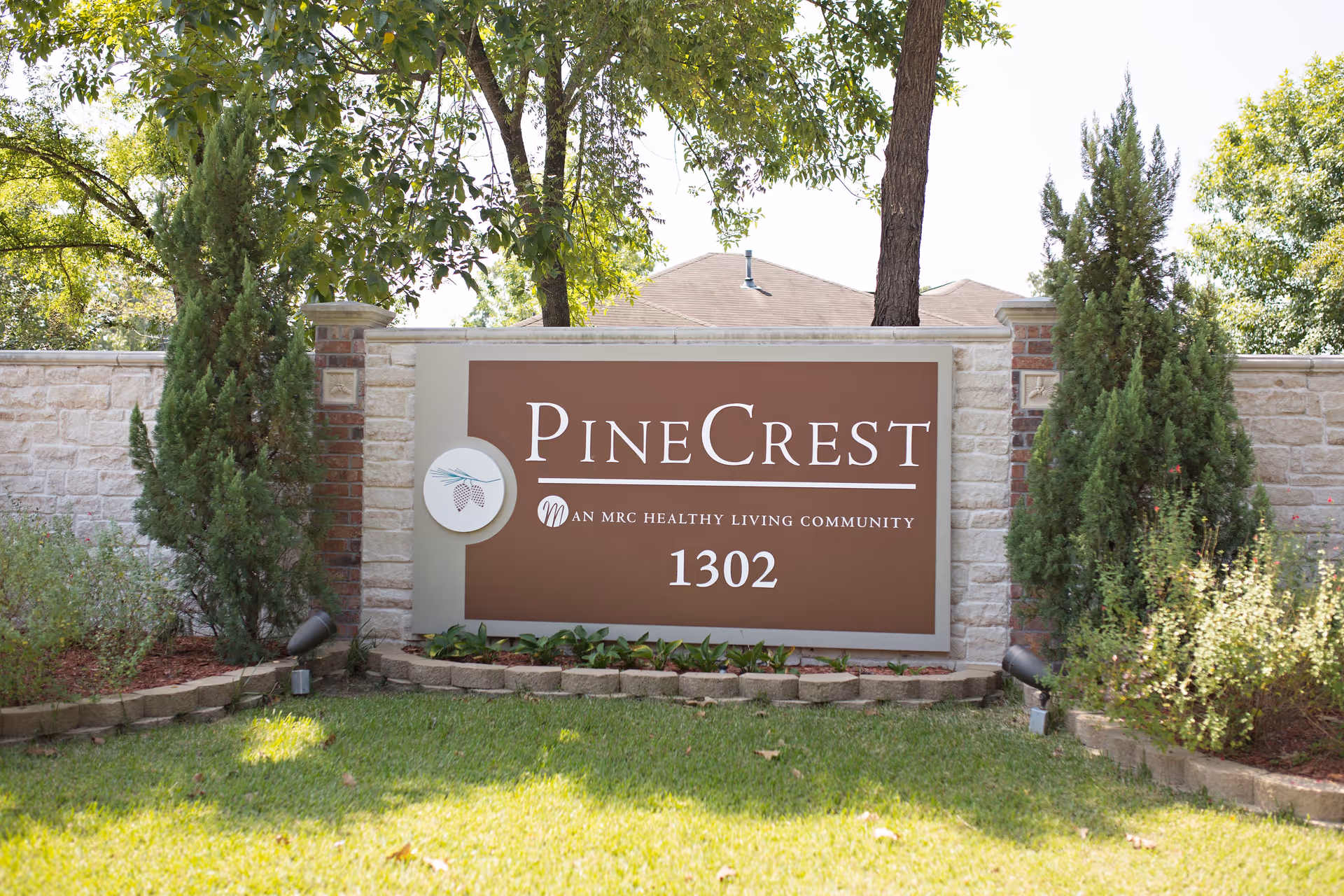 Stone entrance sign reading "PineCrest 1302" surrounded by landscaping, trees, and a lawn.
