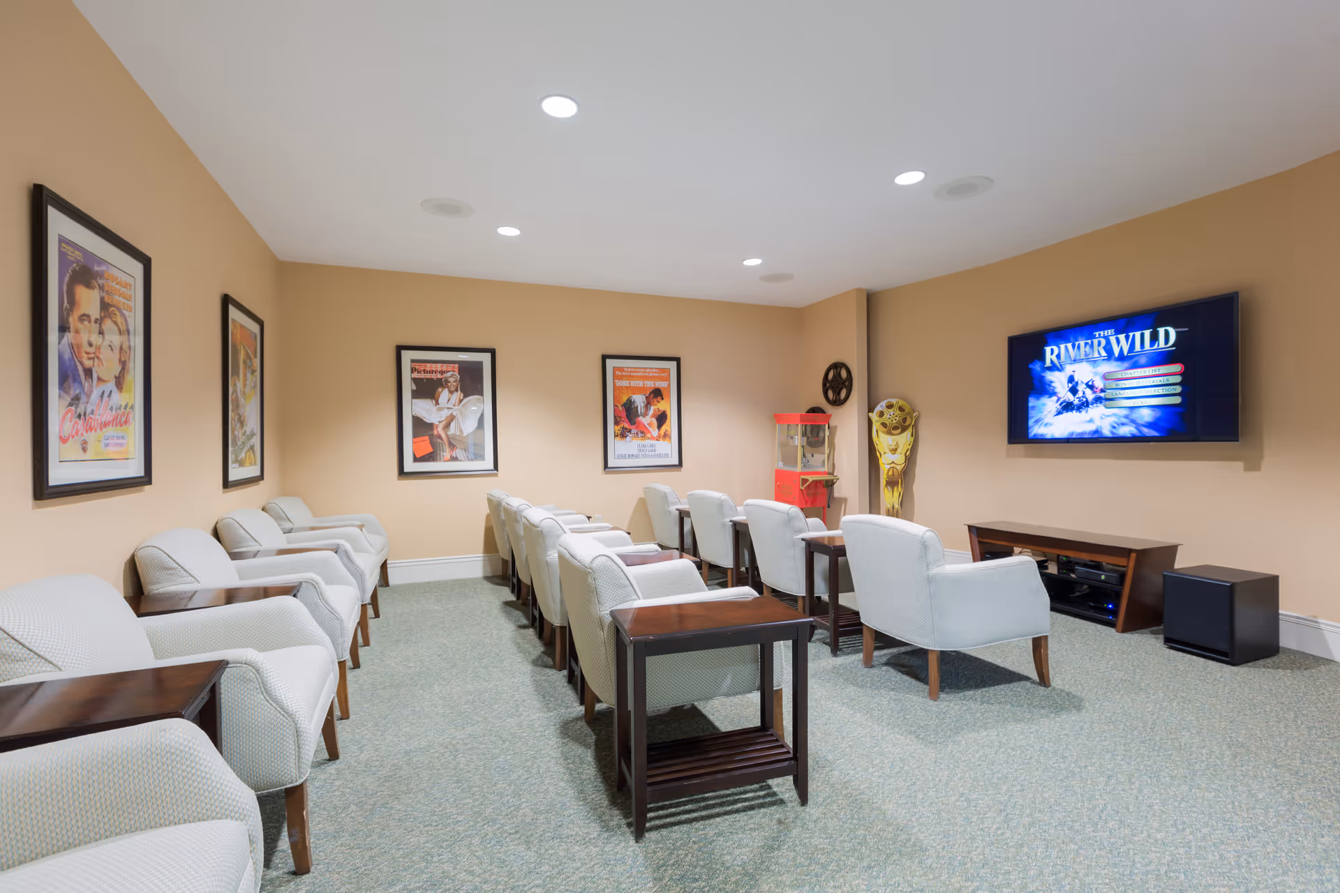 A small movie theater room with two rows of beige armchairs facing a wall-mounted flat screen TV displaying the movie 'The River Wild'. The room has beige walls adorned with framed classic movie posters and a popcorn machine in the corner. There are small wooden side tables between some chairs and a carpeted floor.