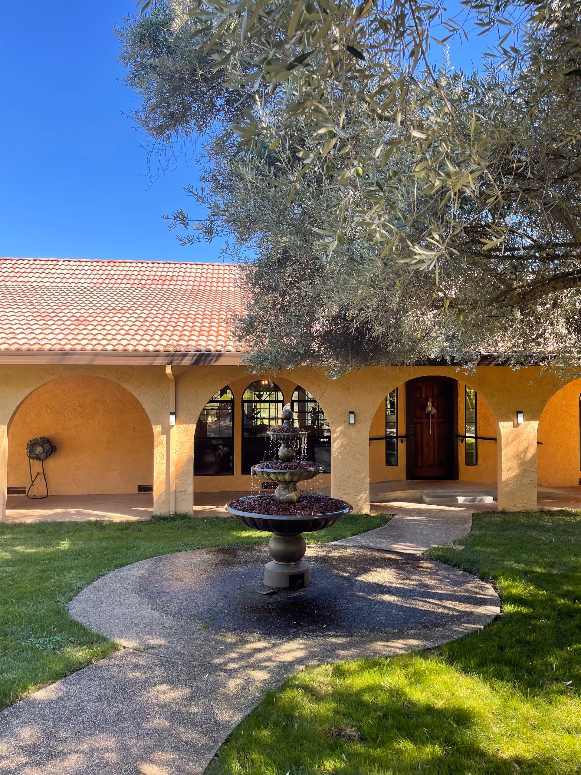 Courtyard with a tiered fountain in front of a stucco building with arched walkways and a red tile roof beneath a large tree.
