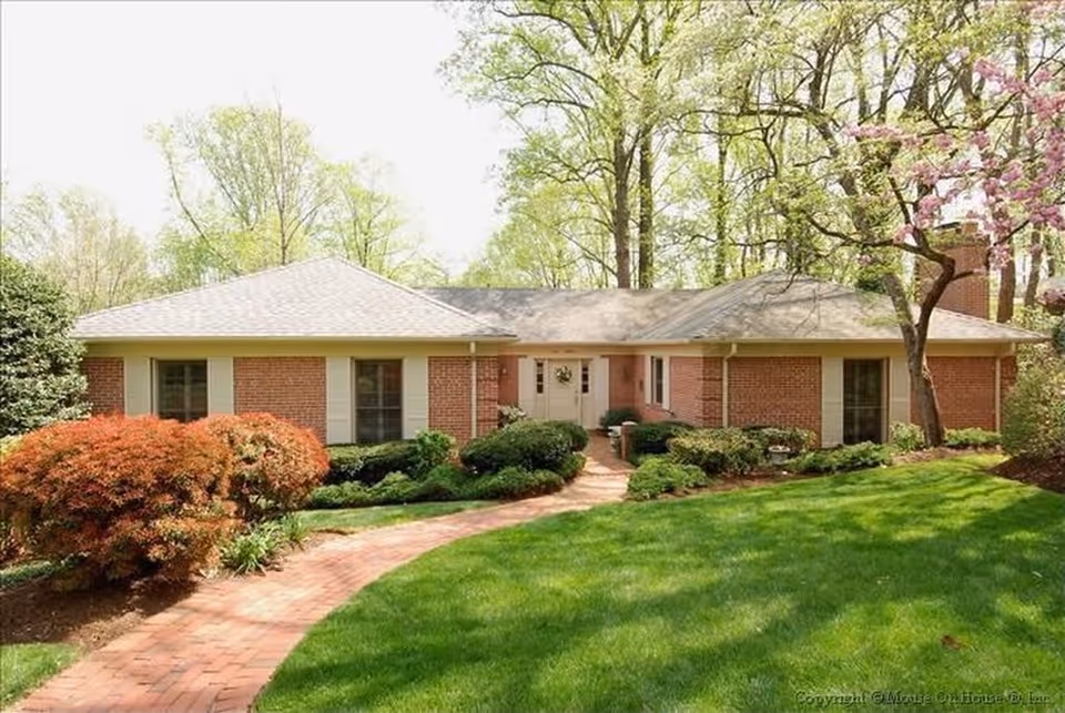 Single-story brick building with a gray shingled roof surrounded by green grass, bushes, and trees, with a curved brick walkway leading to the front door.
