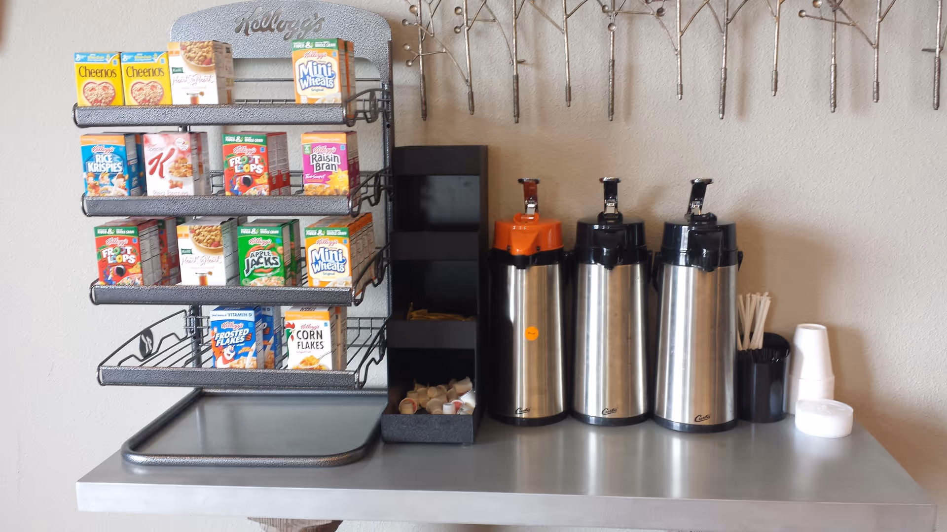 A breakfast station with a rack holding various small boxes of Kellogg's cereals including Cheerios, Mini Wheats, Rice Krispies, Special K, Froot Loops, Raisin Bran, Apple Jacks, Frosted Flakes, and Corn Flakes. Next to the cereal rack are three stainless steel airpots for coffee or hot beverages, a black container holding creamers and sugar packets, a cup with stirrers, and a stack of white disposable cups on a gray countertop. The wall behind has a decorative metal wall art piece.