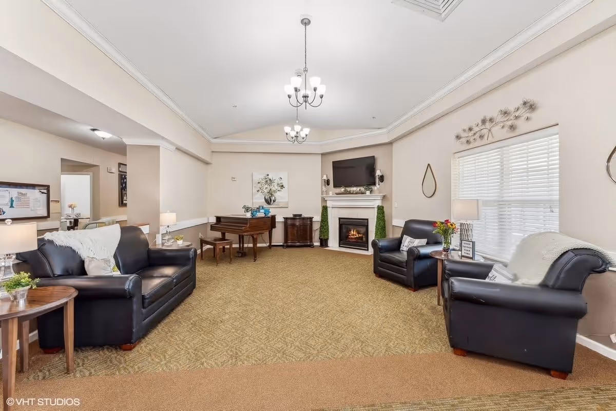A cozy living room with beige walls and carpeted floor featuring three black leather armchairs and a sofa arranged around a fireplace with a mounted TV above it. There is a wooden piano with a bench against the far wall, a small round wooden side table with a lamp and flowers, and decorative wall art including a mirror and metal wall sculpture. The room is well-lit with ceiling chandeliers and natural light from a window with white blinds.