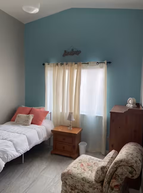 A cozy bedroom with a single bed on the left side, adorned with white bedding and pink and white pillows. In the center, there is a wooden nightstand with a small lamp and a white trash bin underneath. A window with sheer cream curtains is in the background, and above it hangs a decorative sign that reads 'Sunshine'. On the right side, there is a wooden dresser with a clock on top and a floral upholstered armchair in the foreground.