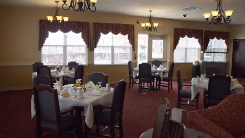 A dining room with multiple round tables covered with white tablecloths, each set with napkins, glasses, and condiments. The room has large windows with red curtains, red carpet, and several chandeliers hanging from the ceiling. There are dark wooden chairs around the tables and a red upholstered sofa in the foreground.